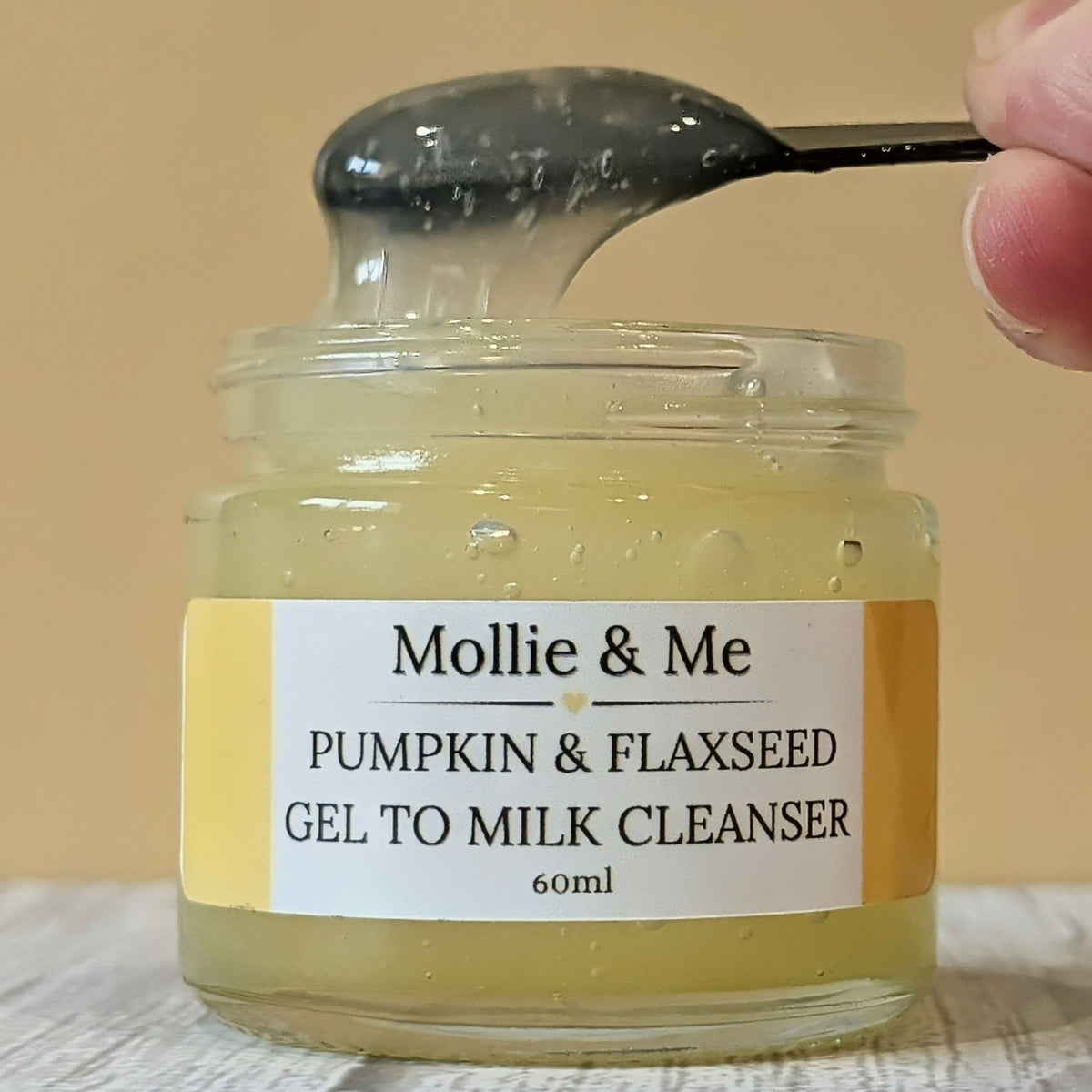 A jar of Mollie & Me gel to milk organic cleanser with a yellow and white label and an applicator lifting some product out of the open jar, showing its thick gel texture and golden colour.