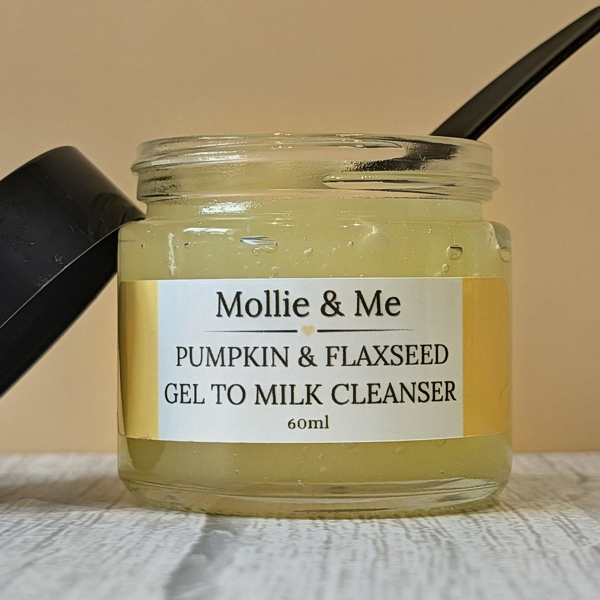Mollie & Me Organic Natural Cleanser for dry and mature skin with Pumpkin & Flaxseed. Gel to Milk Cleanser with a black lid resting beside it on a light wood surface against a beige background.