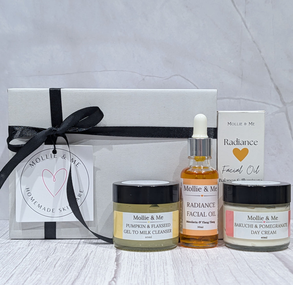 A natural skincare routine set for mature, dry and sensitive skin from Mollie & Me, featuring a white gift box with a black ribbon, a facial oil bottle, a cleanser jar, and a day cream jar, arranged on a light wood surface against a gray marble background. Made in small batches in Kent.
